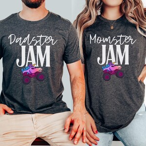 May include: Two dark gray t-shirts with the phrases "Dadster JAM" and "Momster JAM" in white script. Each shirt features a cartoon monster truck with a unicorn head in pink, purple, and blue. The shirts are short-sleeved.