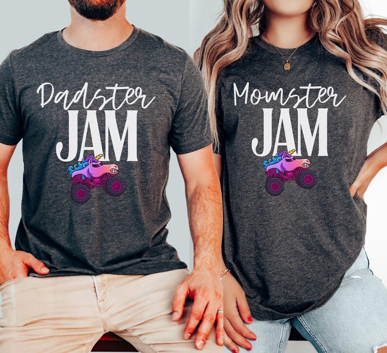 May include: Two dark gray t-shirts with the phrases "Dadster JAM" and "Momster JAM" in white script. Each shirt features a cartoon monster truck with a unicorn head in pink, purple, and blue. The shirts are short-sleeved.