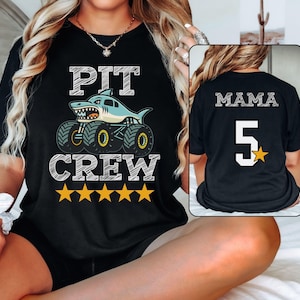 Peut inclure: T-shirt noir avec un motif de camion monstre requin et les mots "PIT CREW". Cinq étoiles jaunes sont en dessous du texte. Le dos du t-shirt porte le mot "MAMA", le chiffre "5" et une étoile jaune.