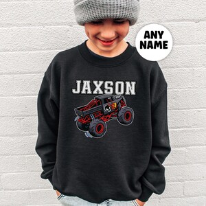 以下が含まれることがあります： 白いブロック文字で「JAXSON」という名前と、赤と黒のディテールが施された漫画のモンスタートラックが特徴の黒いスウェットシャツ。 トラックは大きな赤いホイールとスカルグラフィックを備えています。