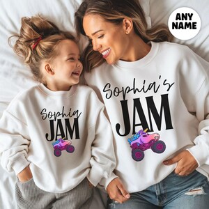 May include: Two white sweatshirts with the text "Sophia's JAM" in black script and block letters. A colorful monster truck graphic is below the text. The sweatshirts are worn by a mother and daughter, who are smiling.