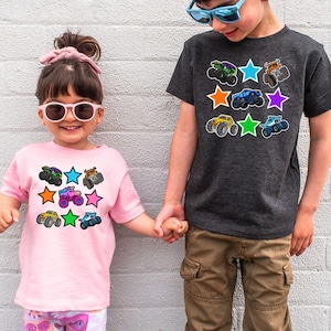 May include: Two children wearing t-shirts with colorful monster truck graphics. The pink shirt has a design of monster trucks and stars. The gray shirt also features monster trucks and stars. Both shirts are short-sleeved.