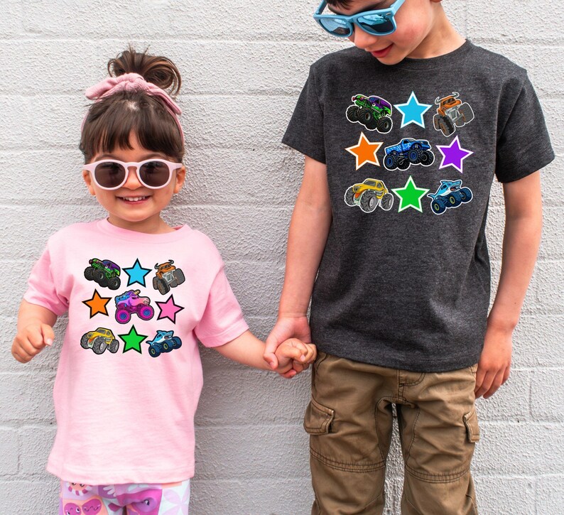 May include: Two children wearing t-shirts with colorful monster truck graphics. The pink shirt has a design of monster trucks and stars. The gray shirt also features monster trucks and stars. Both shirts are short-sleeved.