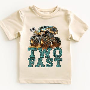 Pictuered is a natural colored t-shirt which says TWO FAST in blue sketch big graphic letters underneath a big monster truck with a checkered flag waving in the breeze behind. there is a custom age number on the side of the truck.