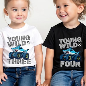 Op de afbeelding: Twee kinderen dragen T-shirts met een haaienmonster truck ontwerp. E&eacute;n is wit en zegt "Young Wild & Three", de andere is zwart en zegt "Young Wild & Four". De shirts hebben blauwe en gele details.