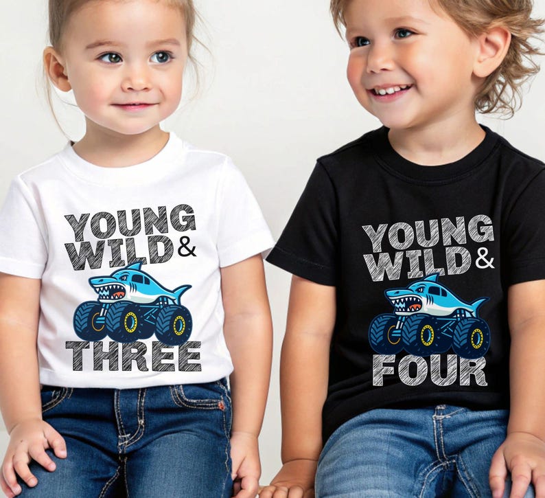 Op de afbeelding: Twee kinderen dragen T-shirts met een haaienmonster truck ontwerp. E&eacute;n is wit en zegt "Young Wild & Three", de andere is zwart en zegt "Young Wild & Four". De shirts hebben blauwe en gele details.
