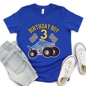 May include: Blue t-shirt with a shark-themed monster truck graphic and the text "BIRTHDAY BOY 3". The design includes checkered flags. The shirt is paired with denim shorts and white sneakers.