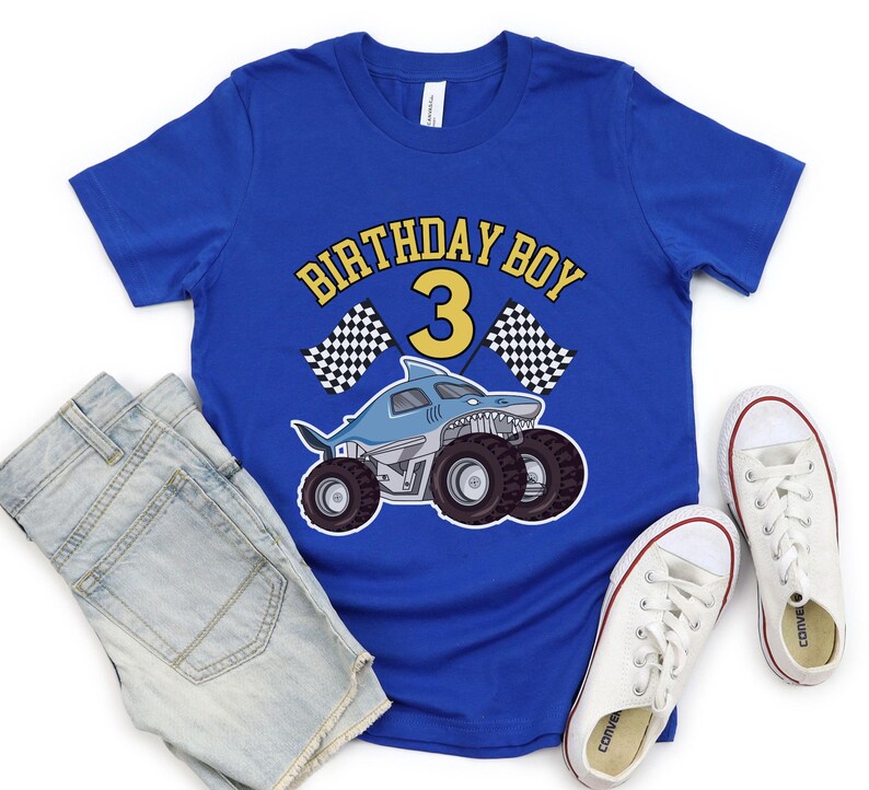 May include: Blue t-shirt with a shark-themed monster truck graphic and the text "BIRTHDAY BOY 3". The design includes checkered flags. The shirt is paired with denim shorts and white sneakers.