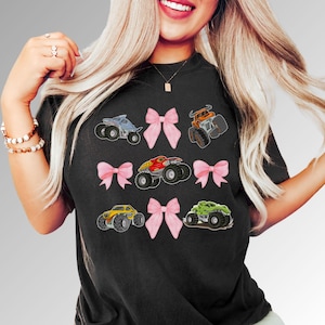 K&ouml;nnte beinhalten: Schwarzes T-Shirt mit farbenfrohen Monstertruck-Grafiken und rosa Schleifenakzenten. Die Trucks sind in verschiedenen Farben, darunter Blau, Orange, Rot, Gelb und Gr&uuml;n. Ein lustiges und verspieltes Design.