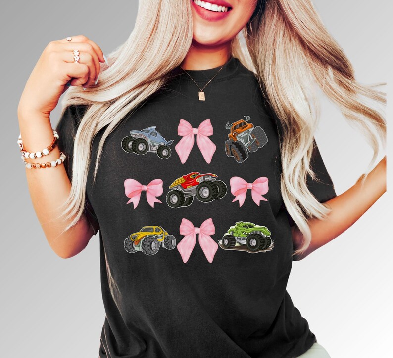 K&ouml;nnte beinhalten: Schwarzes T-Shirt mit farbenfrohen Monstertruck-Grafiken und rosa Schleifenakzenten. Die Trucks sind in verschiedenen Farben, darunter Blau, Orange, Rot, Gelb und Gr&uuml;n. Ein lustiges und verspieltes Design.