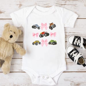 Op de afbeelding: Witte babybody met geborduurde monstertruckontwerpen in blauw, oranje, rood, geel en groen. Roze strikken zijn geborduurd tussen de trucks. Een teddybeer en zwart-witte babyschoenen liggen ernaast.