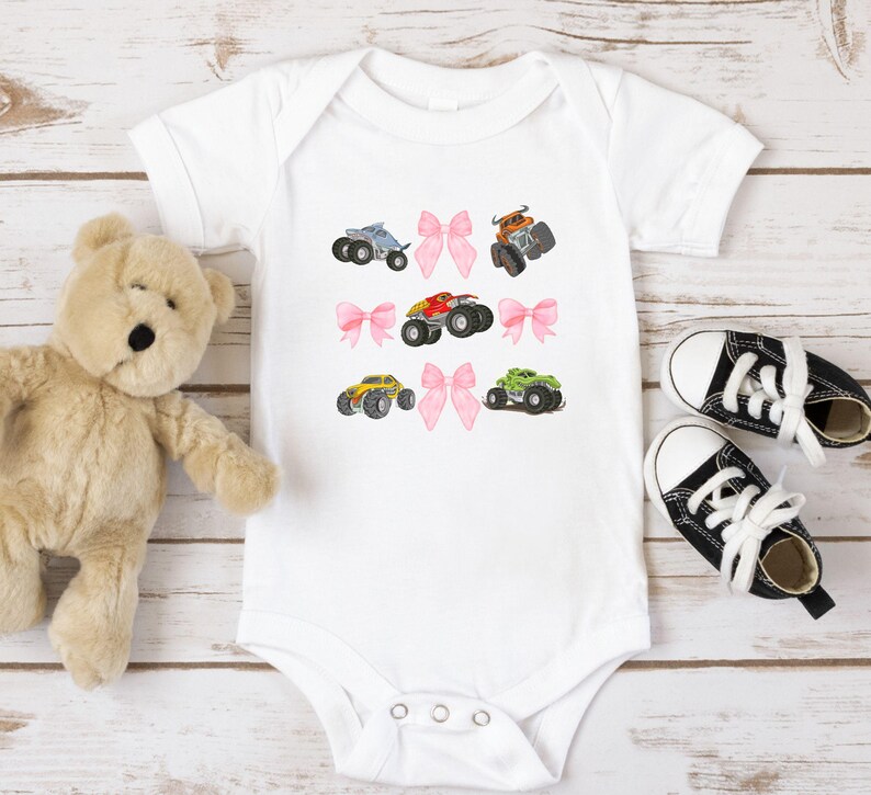 Op de afbeelding: Witte babybody met geborduurde monstertruckontwerpen in blauw, oranje, rood, geel en groen. Roze strikken zijn geborduurd tussen de trucks. Een teddybeer en zwart-witte babyschoenen liggen ernaast.