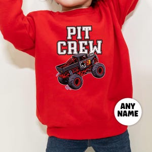 以下が含まれることがあります： 「PIT CREW」の文字が白のブロック体で書かれた赤いスウェットシャツ。黒と赤のモンスタートラックのグラフィックが上にあります。「ANY NAME」という文字が入った白い円が右下にあります。
