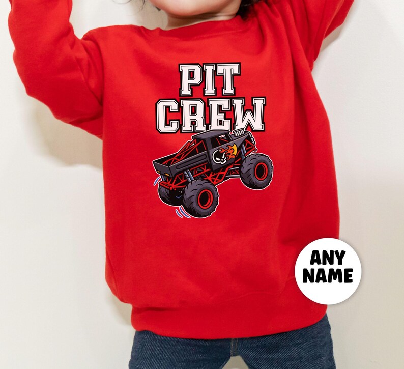 以下が含まれることがあります： 「PIT CREW」の文字が白のブロック体で書かれた赤いスウェットシャツ。黒と赤のモンスタートラックのグラフィックが上にあります。「ANY NAME」という文字が入った白い円が右下にあります。