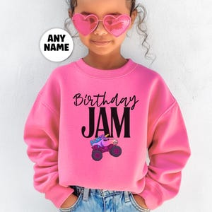 May include: A pink sweatshirt with the words "Birthday JAM" in black script and block letters. A cartoon monster truck with a unicorn on top is below the text. A white circle with the words "ANY NAME" is in the upper left.