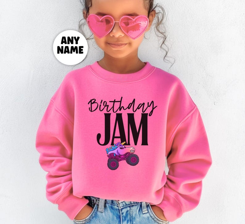 May include: A pink sweatshirt with the words "Birthday JAM" in black script and block letters. A cartoon monster truck with a unicorn on top is below the text. A white circle with the words "ANY NAME" is in the upper left.