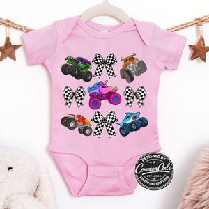 May include: A light pink baby bodysuit featuring colorful monster truck graphics and checkered bows. The trucks include a green monster truck, an orange monster truck, a red monster truck, a blue shark truck, and a unicorn truck. The text "DESIGNED BY COMMON CODE EST. 2020 FOR YOU AND YOUR CREW" is printed on the bottom right.