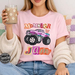 Puede incluir: Camiseta rosa claro con las palabras "Momster Jam" en letras coloridas y cuadradas. Un gr&aacute;fico de un cami&oacute;n monstruo rosa est&aacute; centrado debajo del texto. La camiseta tiene mangas cortas y cuello redondo.