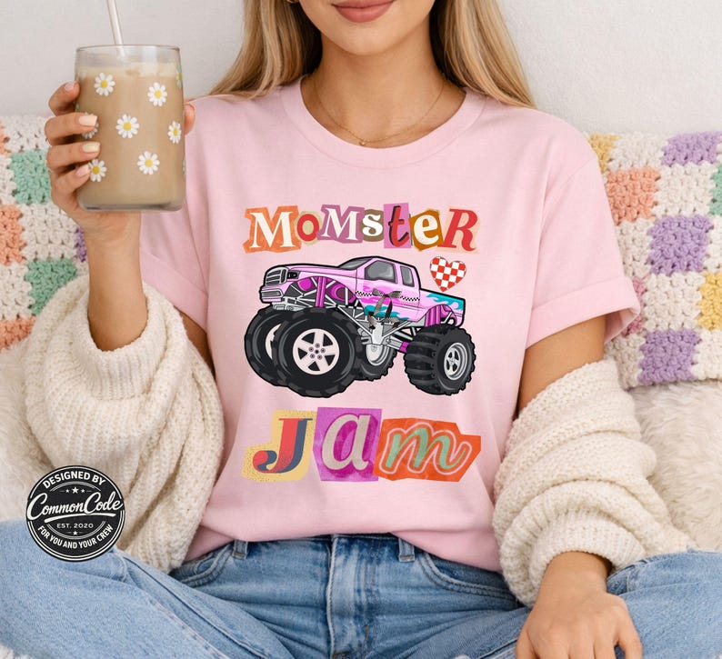 Puede incluir: Camiseta rosa claro con las palabras "Momster Jam" en letras coloridas y cuadradas. Un gr&aacute;fico de un cami&oacute;n monstruo rosa est&aacute; centrado debajo del texto. La camiseta tiene mangas cortas y cuello redondo.