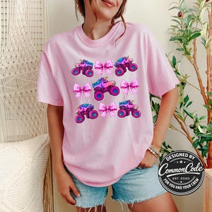 May include: A light pink t-shirt with a repeating pattern of cartoon monster trucks with unicorn horns and pink bows. The shirt has a crew neck and short sleeves. The text "DESIGNED BY Common Code FOR YOU AND YOUR CREW EST. 2020" is on the bottom right.