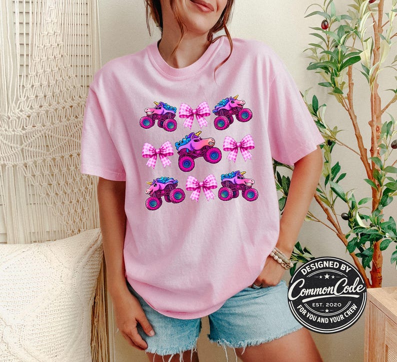 May include: A light pink t-shirt with a repeating pattern of cartoon monster trucks with unicorn horns and pink bows. The shirt has a crew neck and short sleeves. The text "DESIGNED BY Common Code FOR YOU AND YOUR CREW EST. 2020" is on the bottom right.