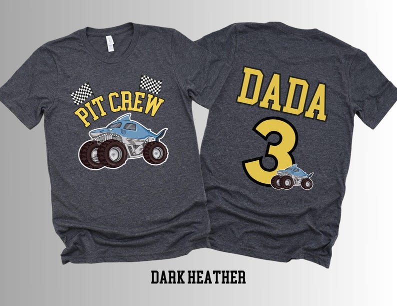 Two dark heather gray t-shirts. The front of one shirt has a blue shark monster truck with the varsity text 'Pit Crew' above it and 2 checkered flags. The back of the other shirt has the college varsity text 'Dada' in yellow with the number '3' below it with a small version of the shark truck.