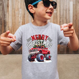 May include: A gray t-shirt featuring a red monster truck with large blue tires and the words "MERRY CHRISTMAS" in a festive design. The shirt has a classic crew neck and short sleeves.