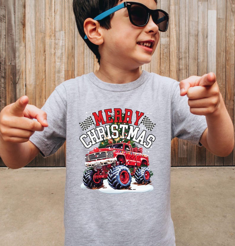 May include: A gray t-shirt featuring a red monster truck with large blue tires and the words "MERRY CHRISTMAS" in a festive design. The shirt has a classic crew neck and short sleeves.