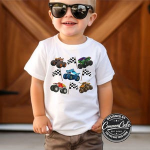 May include: White t-shirt featuring colorful monster truck graphics and checkered flags. The shirt has a crew neck and short sleeves. The design includes various monster trucks in different colors, such as orange, green, and blue.