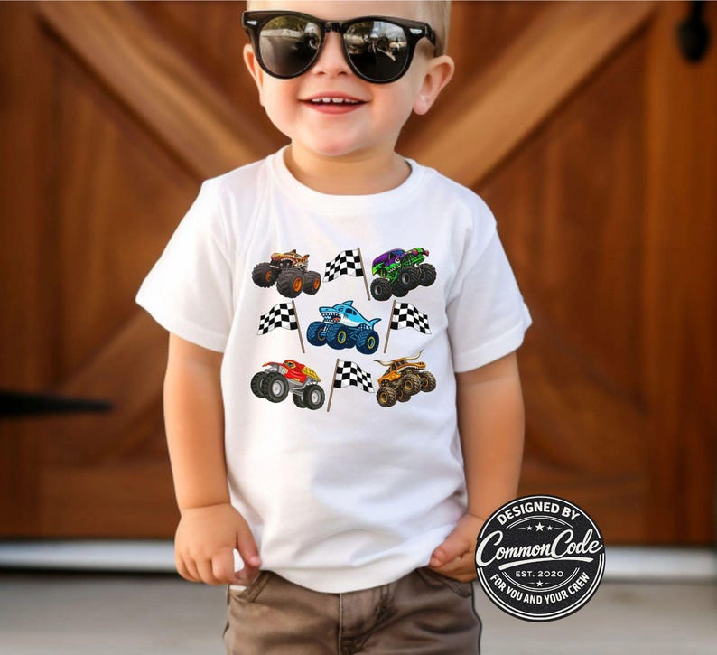May include: White t-shirt featuring colorful monster truck graphics and checkered flags. The shirt has a crew neck and short sleeves. The design includes various monster trucks in different colors, such as orange, green, and blue.