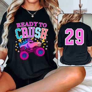 Pode incluir: Camiseta preta com a frase "READY TO CRUSH" acima de um desenho animado de um monster truck com uma cabe&ccedil;a de unic&oacute;rnio. O caminh&atilde;o &eacute; roxo, rosa e azul. A parte de tr&aacute;s da camisa tem o n&uacute;mero "29" em rosa.