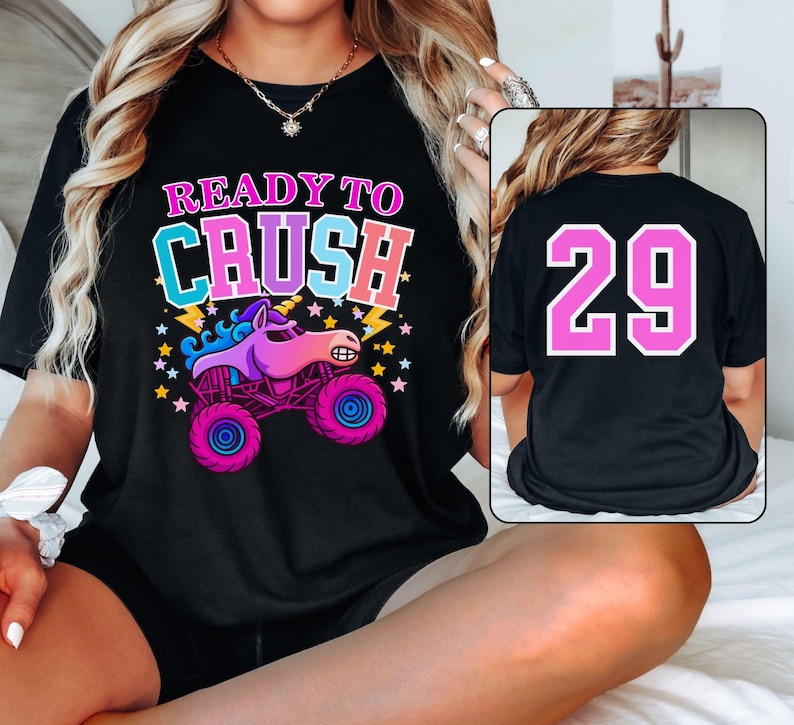 Pode incluir: Camiseta preta com a frase "READY TO CRUSH" acima de um desenho animado de um monster truck com uma cabe&ccedil;a de unic&oacute;rnio. O caminh&atilde;o &eacute; roxo, rosa e azul. A parte de tr&aacute;s da camisa tem o n&uacute;mero "29" em rosa.