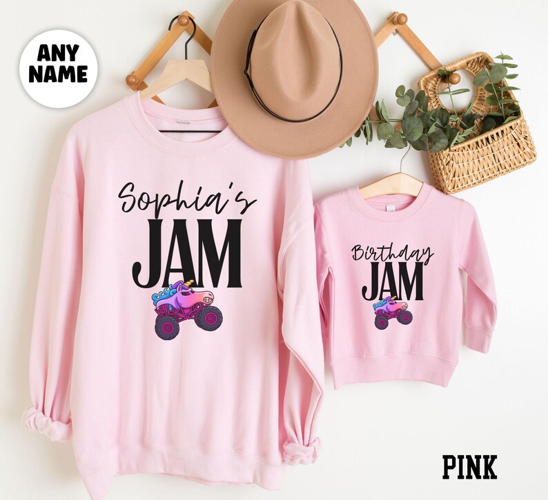 May include: Pink sweatshirts with the text "Sophia's JAM" and "Birthday JAM". Each sweatshirt features a colorful monster truck graphic. A tan hat and a small basket with greenery are also visible.