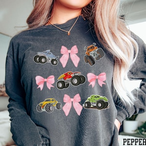 Pictured is a woman wearing a dark gray long-sleeve T-shirt featuring colorful monster truck graphics and pink bows, 9 total in 3 rows of 3. The monster trucks are in various colors: blue, orange, red, yellow, and green. The word 'PEPPER' is printed in white at the bottom right corner, denoting the color of the shirt