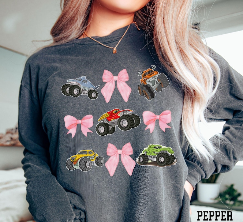 Pictured is a woman wearing a dark gray long-sleeve T-shirt featuring colorful monster truck graphics and pink bows, 9 total in 3 rows of 3. The monster trucks are in various colors: blue, orange, red, yellow, and green. The word 'PEPPER' is printed in white at the bottom right corner, denoting the color of the shirt