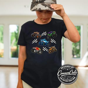 May include: Black t-shirt featuring colorful monster truck graphics with checkered flags. The design includes various monster trucks in orange, green, blue, and red. The shirt has the text "DESIGNED BY Common Code" and "FOR YOU AND YOUR CREW".