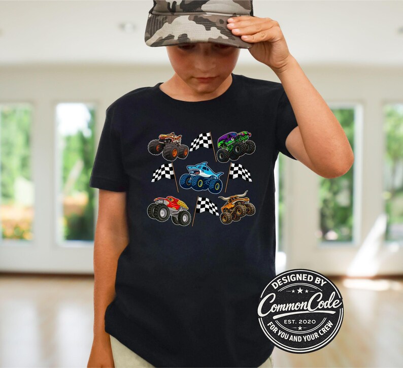May include: Black t-shirt featuring colorful monster truck graphics with checkered flags. The design includes various monster trucks in orange, green, blue, and red. The shirt has the text "DESIGNED BY Common Code" and "FOR YOU AND YOUR CREW".