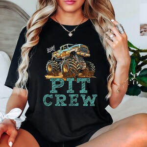 Pictured is a woman wearing a black t-shirt which says PIT CREW in blue big graphic letters underneath a monster truck with a custom number on the side and a black and white checkered flag waving behind