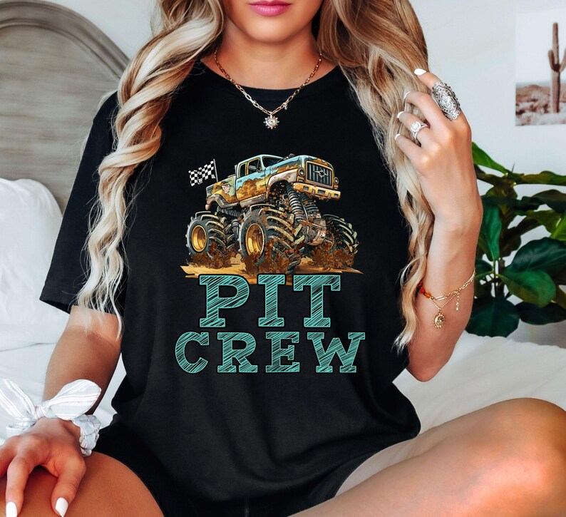 Pictured is a woman wearing a black t-shirt which says PIT CREW in blue big graphic letters underneath a monster truck with a custom number on the side and a black and white checkered flag waving behind