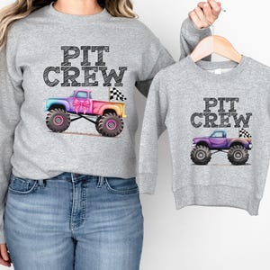 Puede incluir: Dos sudaderas grises con el texto "PIT CREW". Una sudadera presenta un cami&oacute;n monstruo colorido con una bandera a cuadros y un lazo rosa. La otra sudadera tiene un cami&oacute;n monstruo morado m&aacute;s peque&ntilde;o con una bandera a cuadros.