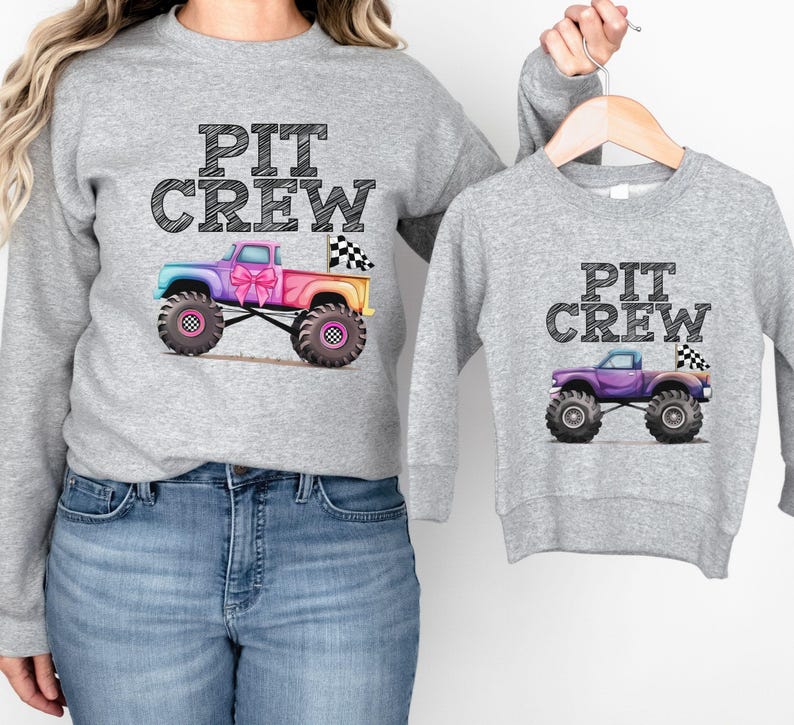 Puede incluir: Dos sudaderas grises con el texto "PIT CREW". Una sudadera presenta un cami&oacute;n monstruo colorido con una bandera a cuadros y un lazo rosa. La otra sudadera tiene un cami&oacute;n monstruo morado m&aacute;s peque&ntilde;o con una bandera a cuadros.