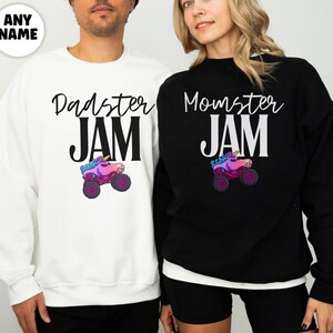 May include: White and black sweatshirts with the text "Dadster JAM" and "Momster JAM" in a script font. Each sweatshirt features a colorful monster truck graphic. The sweatshirts are worn by two people.