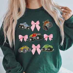 May include: Dark green sweatshirt featuring colorful monster truck graphics and pink bows. The monster trucks are in various colors, including blue, orange, red, yellow, and green. The bows are a soft pink color.