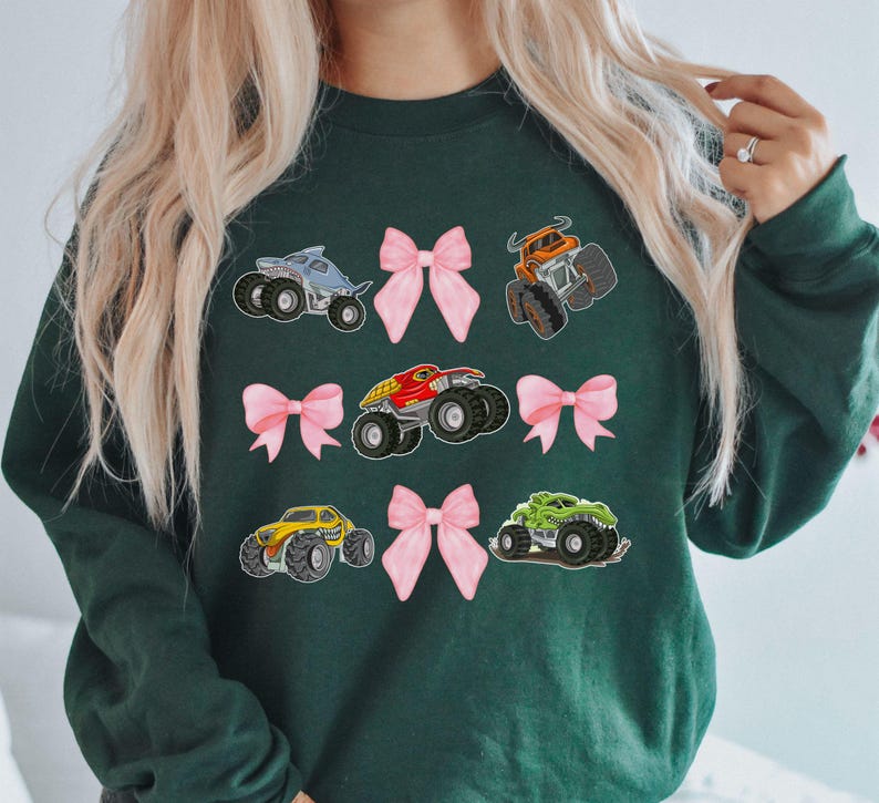 May include: Dark green sweatshirt featuring colorful monster truck graphics and pink bows. The monster trucks are in various colors, including blue, orange, red, yellow, and green. The bows are a soft pink color.