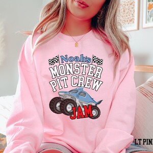 May include: A light pink sweatshirt with the text "Noah's Monster Pit Crew Jam" and a shark-themed monster truck graphic. The design includes black and white checkered flags and bold red lettering.
