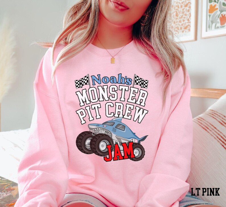 May include: A light pink sweatshirt with the text "Noah's Monster Pit Crew Jam" and a shark-themed monster truck graphic. The design includes black and white checkered flags and bold red lettering.