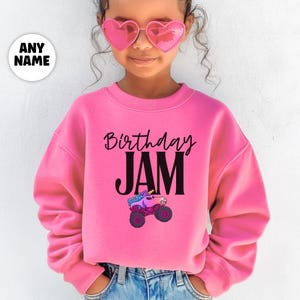 May include: A pink sweatshirt with the words "Birthday JAM" in black script and block letters. A cartoon monster truck with a unicorn head is printed below the text. The sweatshirt is worn by a person wearing pink heart-shaped sunglasses.
