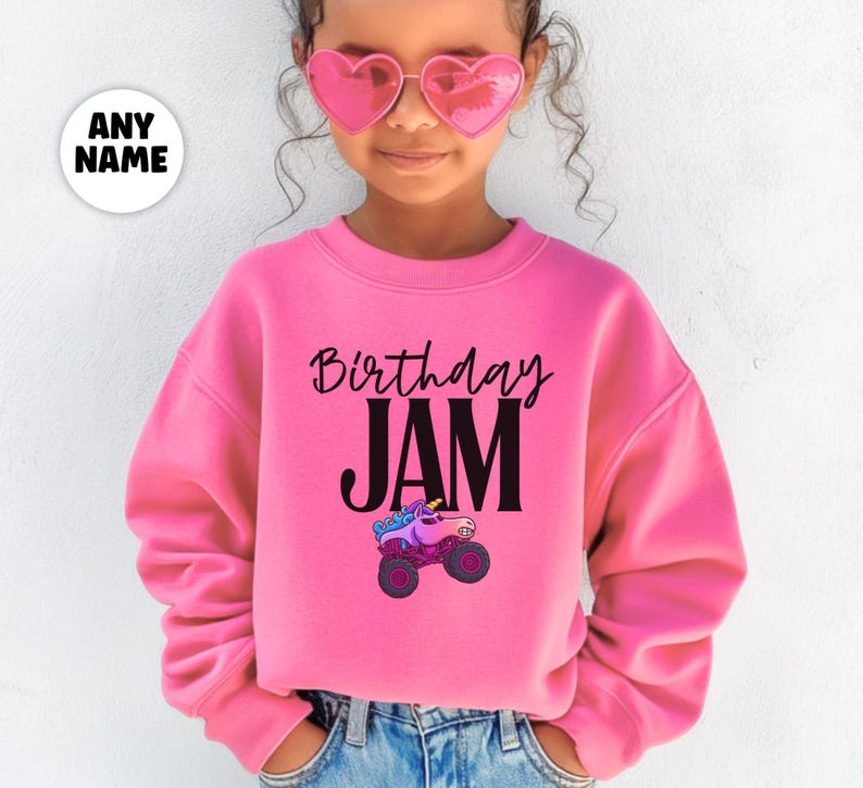May include: A pink sweatshirt with the words "Birthday JAM" in black script and block letters. A cartoon monster truck with a unicorn head is printed below the text. The sweatshirt is worn by a person wearing pink heart-shaped sunglasses.