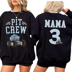 Op de afbeelding: Zwart sweatshirt met de tekst "PIT CREW" en een cartoon monstertruck met een haaienbek. De achterkant van het sweatshirt zegt "MAMA 3" met een bijpassende monstertruckafbeelding. De sweatshirt heeft een witte ronde hals.
