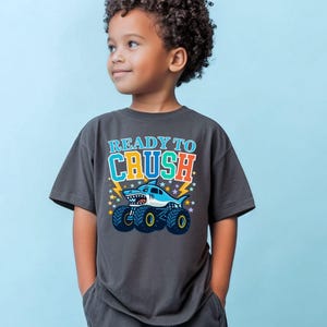 May include: Dark gray t-shirt with the text "READY TO CRUSH" above a cartoon shark monster truck. The shark is light blue with a white belly and a toothy grin. The truck has large black tires with yellow rims.